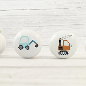 May include: Four white ceramic knobs with different construction vehicle designs. The knobs feature a blue excavator, an orange crane, a yellow stop sign, and a brown crane.