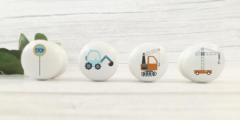 May include: Four white ceramic knobs with different construction vehicle designs. The knobs feature a blue excavator, an orange crane, a yellow stop sign, and a brown crane.