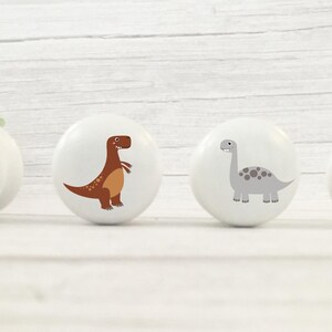 Four white solid Beech furniture knobs with colourful dinosaur designs. The dinosaurs include an orange Triceratops, a orange/beige Stegosaurus, a brown T Rex and a grey Brachiosaurus. The drawer handles have a smooth glossy finish and are shown on a pale wood background.