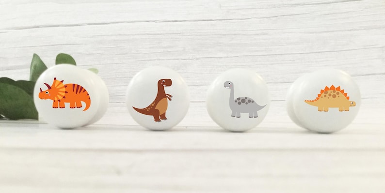 Four white solid Beech furniture knobs with colourful dinosaur designs. The dinosaurs include an orange Triceratops, a orange/beige Stegosaurus, a brown T Rex and a grey Brachiosaurus. The drawer handles have a smooth glossy finish and are shown on a pale wood background.