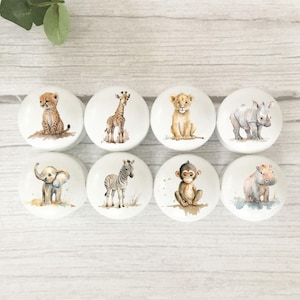 May include: Eight white ceramic cabinet knobs with watercolour illustrations of safari animals. The knobs feature a cheetah, giraffe, lion cub, rhino, elephant, zebra, monkey, and hippo. Each knob is approximately 3.8 cm in diameter.