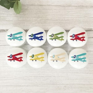 May include: Eight white cabinet knobs with biplane designs in various colours. Each knob features a different coloured airplane, including blue, green, red, and yellow. The knobs are round and have a smooth surface, suitable for a child's room or nursery.
