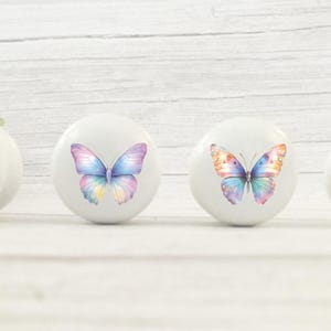 Four white round solid beech furniture knobs each with a pastel rainbow butterfly design. The drawer handles have a glossy finish and are shown on a pale wood background with foliage for decoration.