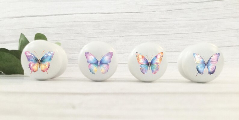 Four white round solid beech furniture knobs each with a pastel rainbow butterfly design. The drawer handles have a glossy finish and are shown on a pale wood background with foliage for decoration.