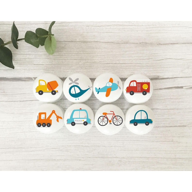 May include: Eight white round knobs with colourful cartoon images of vehicles, including a cement truck, helicopter, aeroplane, fire engine, excavator, police car, bicycle, and car.