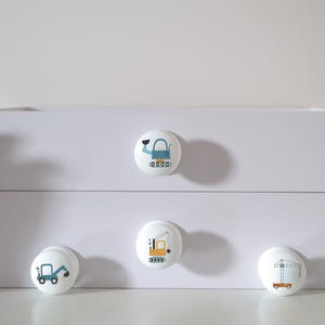 A white chest of drawers with four solid beech furniture knobs featuring construction vehicle designs. The knobs have images of a crane, a digger, an excavator, and a wrecking ball. A succulent plant in a white pot is on the left for decoration.