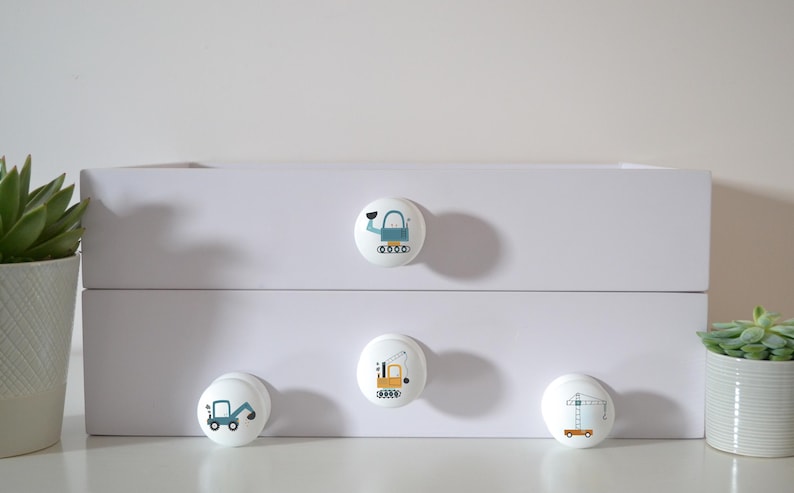 A white chest of drawers with four solid beech furniture knobs featuring construction vehicle designs. The knobs have images of a crane, a digger, an excavator, and a wrecking ball. A succulent plant in a white pot is on the left for decoration.
