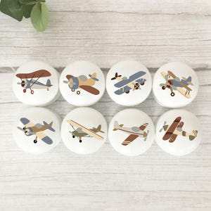 May include: A set of eight round white knobs, each featuring a unique illustration of a vintage aeroplane in shades of brown, blue, and tan. The aeroplanes are depicted in various flight positions, including biplanes and monoplanes, with visible propellers.