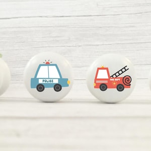 May include: Four white ceramic cabinet knobs with vehicle designs. The knobs feature a blue tractor, a blue police car, a red fire engine, and an orange car. Each knob has a black base and is set against a white wooden background.