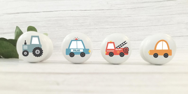 May include: Four white ceramic cabinet knobs with vehicle designs. The knobs feature a blue tractor, a blue police car, a red fire engine, and an orange car. Each knob has a black base and is set against a white wooden background.