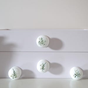 K&ouml;nnte beinhalten: Wei&szlig;er Schrank mit vier runden Schubladengriffen. Jeder Griff zeigt ein botanisches Design aus gr&uuml;nen Bl&auml;ttern und St&auml;ngeln auf wei&szlig;em Hintergrund. Der Schrank wird von Topf-Sukkulenten flankiert.