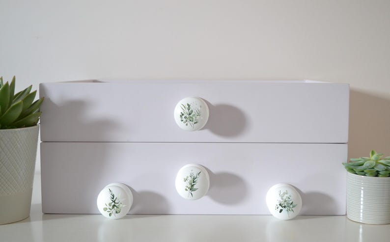 K&ouml;nnte beinhalten: Wei&szlig;er Schrank mit vier runden Schubladengriffen. Jeder Griff zeigt ein botanisches Design aus gr&uuml;nen Bl&auml;ttern und St&auml;ngeln auf wei&szlig;em Hintergrund. Der Schrank wird von Topf-Sukkulenten flankiert.
