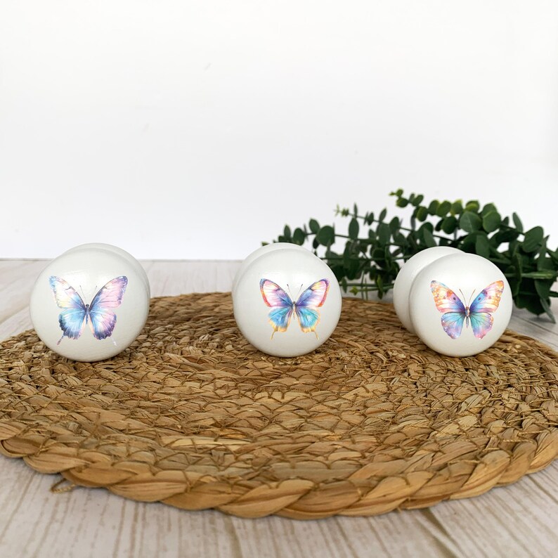 Three white round solid beech furniture knobs each with a rainbow butterfly design in a pastel colour scheme. The cabinet pulls have a glossy finish and are shown on a pale wood background with a wicker mat and foliage for decoration.
