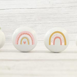 Peut inclure: Quatre boutons en c&eacute;ramique blanche avec des motifs arc-en-ciel. Chaque bouton pr&eacute;sente un arc-en-ciel avec des couleurs rose, jaune et orange, et un petit c&oelig;ur rose au centre. Les arcs-en-ciel sont d&eacute;cor&eacute;s de petits points blancs.