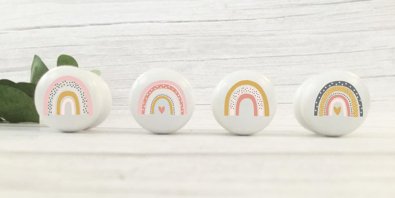 Peut inclure: Quatre boutons en c&eacute;ramique blanche avec des motifs arc-en-ciel. Chaque bouton pr&eacute;sente un arc-en-ciel avec des couleurs rose, jaune et orange, et un petit c&oelig;ur rose au centre. Les arcs-en-ciel sont d&eacute;cor&eacute;s de petits points blancs.