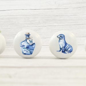 Four white round solid beech furniture knobs each with a different coastal design. The designs include a dolphin ships bottle, sealion and an anchor. The dresser pulls have a glossy finish and are shown on a pale wood background with foliage for decoration.