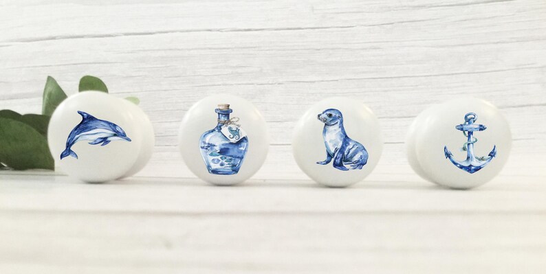 Four white round solid beech furniture knobs each with a different coastal design. The designs include a dolphin ships bottle, sealion and an anchor. The dresser pulls have a glossy finish and are shown on a pale wood background with foliage for decoration.