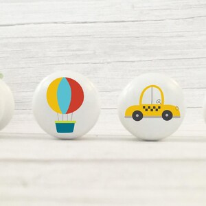 May include: Four white ceramic knobs with different colourful designs. The designs include an ambulance, a hot air balloon, a yellow taxi cab, and a red stop sign.