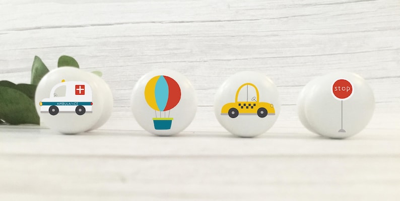 May include: Four white ceramic knobs with different colourful designs. The designs include an ambulance, a hot air balloon, a yellow taxi cab, and a red stop sign.