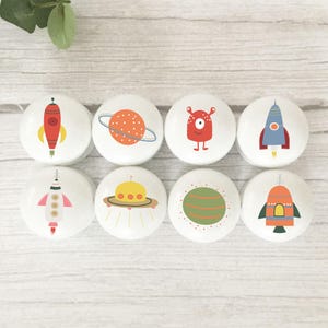 May include: A set of eight round white cabinet knobs featuring colourful space-themed illustrations. The designs include various rockets, a planet with rings, a green planet, a yellow UFO, and a red one-eyed alien character on each individual knob.
