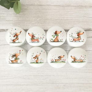 May include: Eight white ceramic cabinet knobs featuring dachshund dog illustrations. Each knob has a unique design of a dachshund with floral accents and butterflies. The knobs are displayed on a light wood surface.