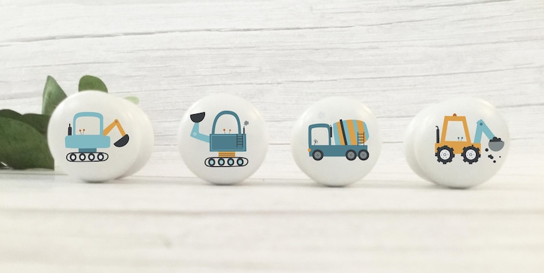 May include: Four ceramic knobs featuring different construction vehicle designs. The knobs are white with blue, yellow and orange accents. The vehicles include a cement mixer, a backhoe, a bulldozer and a dumper truck.