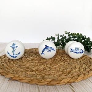 Three white round solid beech furniture knobs each with a different sea themed design. The designs are an anchor, dolphin and a submarine. The cabinet knobs have a glossy finish and are shown on a wicker mat with foliage for decorartion.