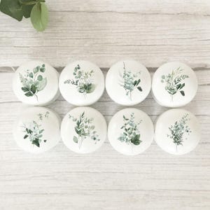 May include: Eight white cabinet knobs with watercolour botanical designs of green leaves and foliage. The knobs are arranged in two rows of four, displaying a variety of plant arrangements.