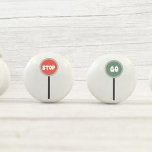 May include: Four white cabinet knobs with traffic-themed designs. One knob features a traffic light, another a red "STOP" sign, a third a green "GO" sign, and the last one reads "BEEP BEEP" in black letters. The knobs are arranged on a white wooden surface.
