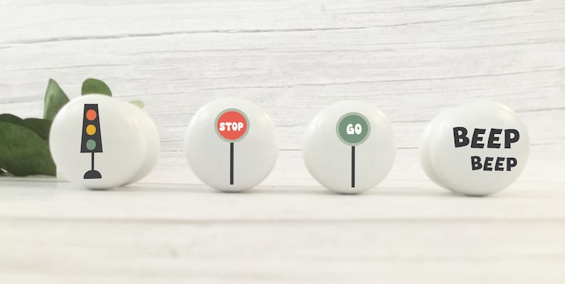 May include: Four white cabinet knobs with traffic-themed designs. One knob features a traffic light, another a red "STOP" sign, a third a green "GO" sign, and the last one reads "BEEP BEEP" in black letters. The knobs are arranged on a white wooden surface.