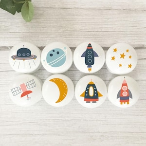 May include: Eight white cabinet knobs with space-themed illustrations. Designs include a UFO, planets, rockets, stars, a satellite, and a crescent moon. These knobs are ideal for adding a playful touch to furniture, with a diameter of approximately 4 cm.