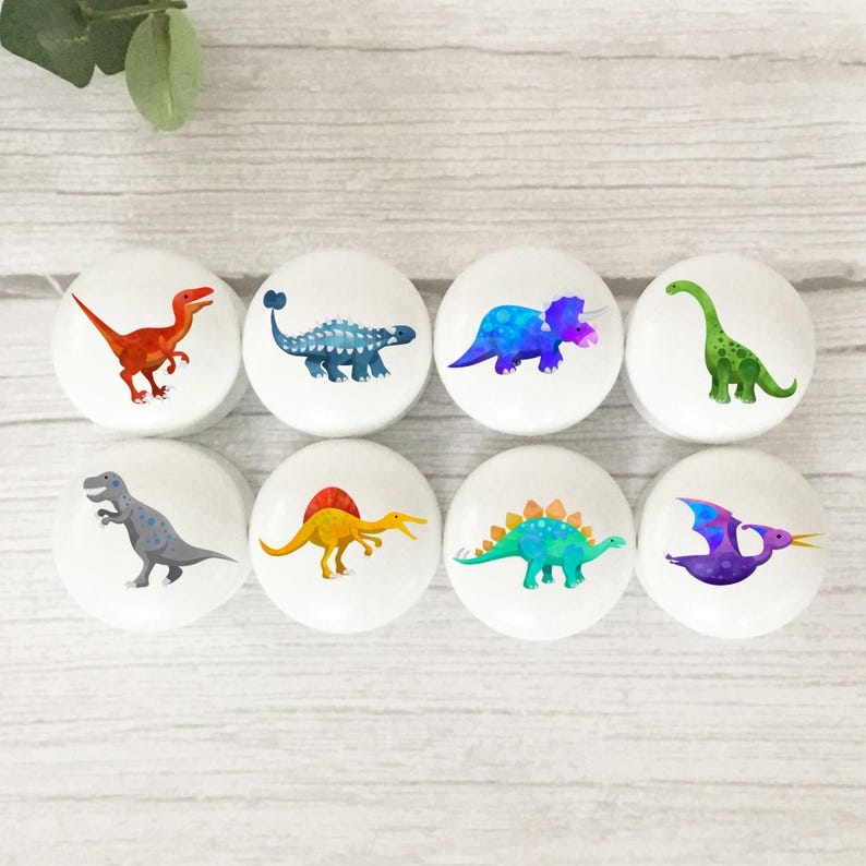 Może przedstawiać: Osiem białych gałek do szafek z kolorowymi ilustracjami dinozaur&oacute;w. Każda gałka przedstawia innego dinozaura w odcieniach czerwieni, niebieskiego, zieleni, ż&oacute;łci i fioletu. Gałki są ułożone w dw&oacute;ch rzędach po cztery.