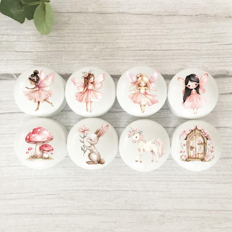 May include: Set of eight white ceramic drawer knobs with whimsical illustrations. The designs include fairies, a unicorn, a bunny, mushrooms, and a small house with a door.