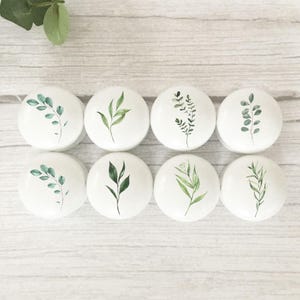 May include: A set of eight round white cabinet knobs, each featuring a unique green botanical illustration of leaves or sprigs. The knobs are arranged in two rows of four, displaying various foliage designs in shades of green on a white surface.