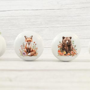 May include: Four white ceramic cabinet knobs with woodland animal designs. The knobs feature a deer, fox, bear, and owl, each surrounded by floral accents. The knobs are round and have a glossy finish.