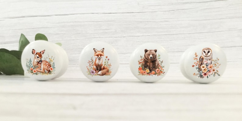 May include: Four white ceramic cabinet knobs with woodland animal designs. The knobs feature a deer, fox, bear, and owl, each surrounded by floral accents. The knobs are round and have a glossy finish.