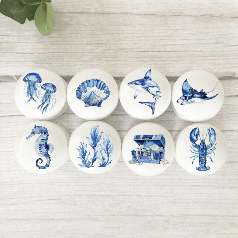 Eight white round solid beech furniture knobs each with a blue nautical themed design. The designs include jellyfish, seashell, shark, stingray, seahorse, seaweed, treasure chest and lobster. The cabinet handles have a glossy finish and are shown on a pale wood background with foliage for decoration.