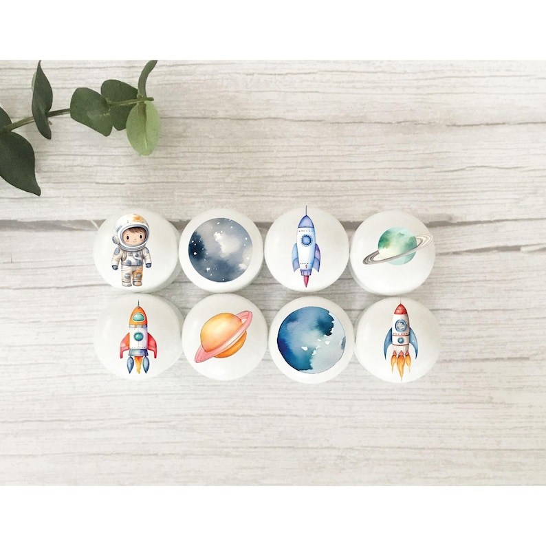 Op de afbeelding: Acht ronde witte keramische knoppen met ruimtethema-illustraties. De ontwerpen omvatten een astronaut, een raket, een planeet en een sterrenhemel.