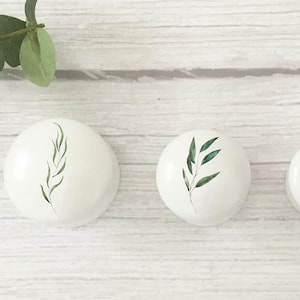 Three solid white beech furniture knobs each with a different leaf design on the front in various shades of green. They are shown in the three sizes available from largest to smallest and are shown on a pale wood  background with foliage for decoration. The cabinet handles have a glossy finish.