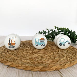 Three white round solid beech furniture knobs each with a different construction vehicle design. They include a crane, truck and excavator. The handles have a glossy finish and are shown on a pale wood background with a wicker mat and foliage for decoration.