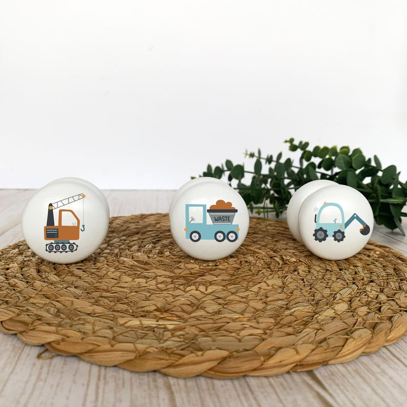 Three white round solid beech furniture knobs each with a different construction vehicle design. They include a crane, truck and excavator. The handles have a glossy finish and are shown on a pale wood background with a wicker mat and foliage for decoration.