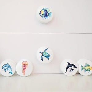 May include: Set of 8 white ceramic drawer knobs with watercolor illustrations of sea creatures, including a fish, turtle, jellyfish, dolphin, orca, fish, crab, and a starfish.