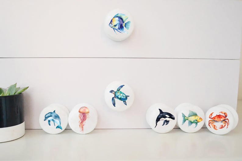 May include: Set of 8 white ceramic drawer knobs with watercolor illustrations of sea creatures, including a fish, turtle, jellyfish, dolphin, orca, fish, crab, and a starfish.