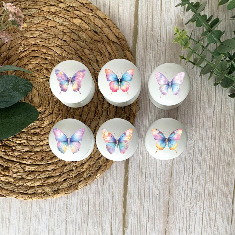 Six white round furniture knobs with a pastel rainbow butterfly designs. The design colours vary but coordinate with different rainbow gradients. The drawer pulls have a glossy finish and are shown on a pale wood background with a wicker mat and foliage for decoration.