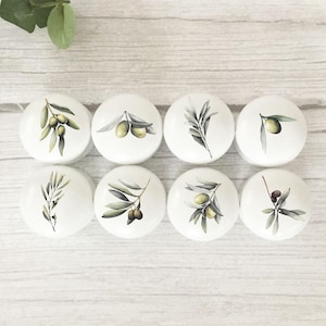 May include: A set of eight round white cabinet knobs, each featuring a unique botanical illustration of olive branches with green and dark purple olives and grey-green leaves, arranged in two rows of four on a light wood grain surface.