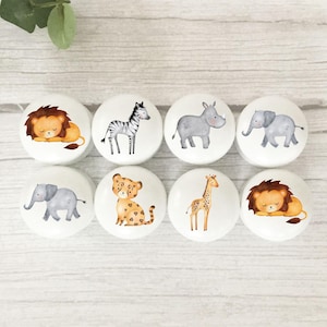 May include: Eight white ceramic cabinet knobs with watercolour animal illustrations. The knobs feature a lion, zebra, rhino, elephant, leopard, giraffe, and two more lions. The knobs are arranged on a light wood surface.