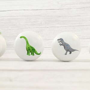 Może przedstawiać: Cztery białe gałki do szafek z ilustracjami dinozaur&oacute;w. Dinozaury są w odcieniach czerwieni, zieleni, szarości i niebieskiego. Gałki są okrągłe i mają błyszczące wykończenie, idealne do pokoju dziecięcego.