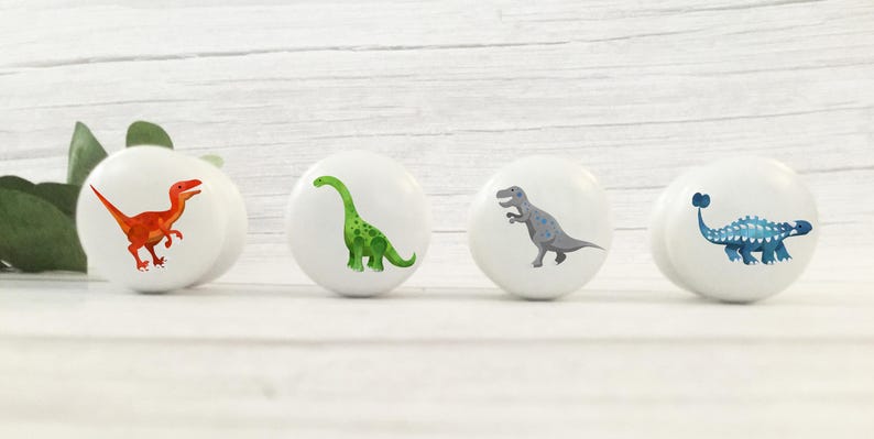 Może przedstawiać: Cztery białe gałki do szafek z ilustracjami dinozaur&oacute;w. Dinozaury są w odcieniach czerwieni, zieleni, szarości i niebieskiego. Gałki są okrągłe i mają błyszczące wykończenie, idealne do pokoju dziecięcego.