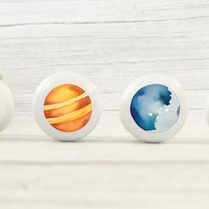 Op de afbeelding: Vier witte keramische knoppen met verschillende ruimtethema-ontwerpen. E&eacute;n knop heeft een cartoon-astronaut, een andere heeft een planeet met oranje en bruine strepen, een derde heeft een blauwe en witte aquarelplaneet en de laatste heeft een rode en witte raket.