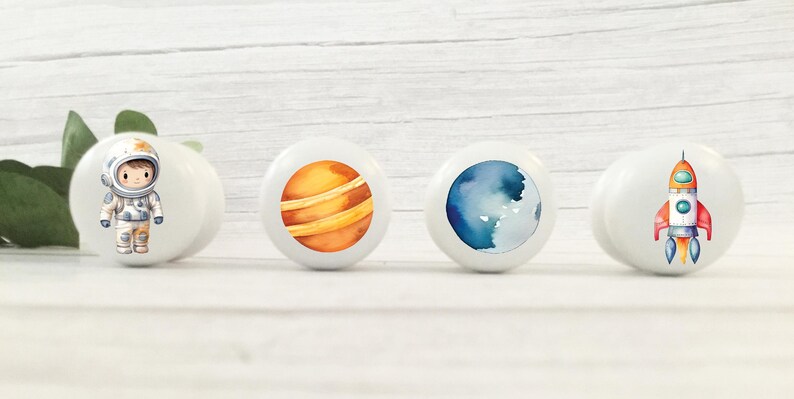Op de afbeelding: Vier witte keramische knoppen met verschillende ruimtethema-ontwerpen. E&eacute;n knop heeft een cartoon-astronaut, een andere heeft een planeet met oranje en bruine strepen, een derde heeft een blauwe en witte aquarelplaneet en de laatste heeft een rode en witte raket.